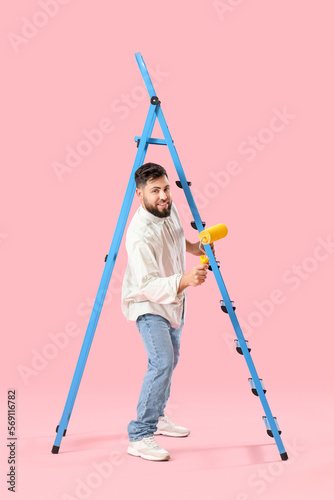 Young man with paint roller and ladder on pink background