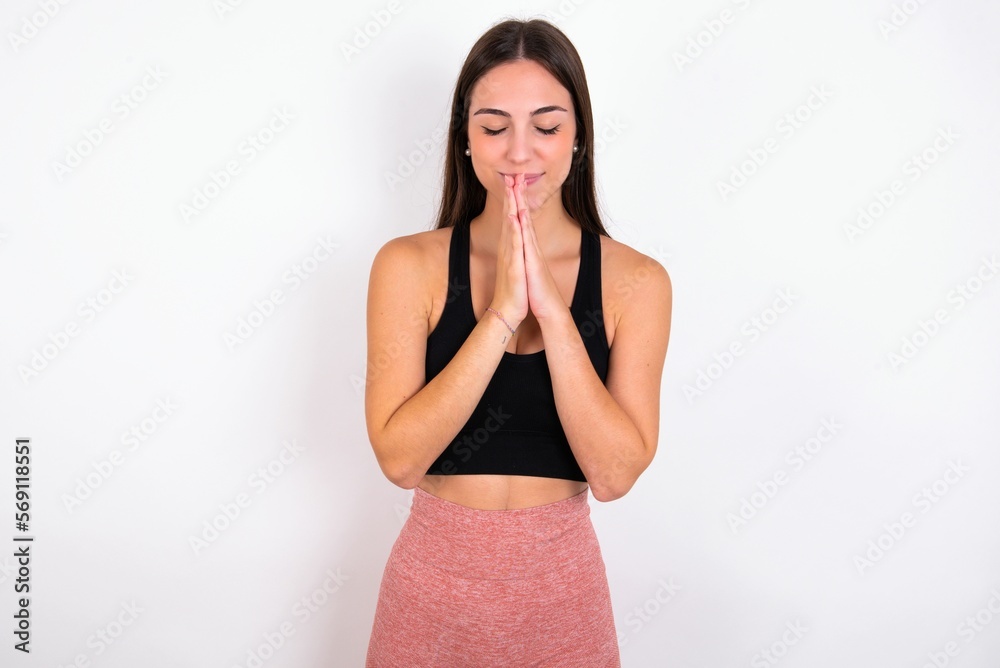 Indoor closeup of young woman wearing sportswear over white studio background practicing yoga and meditation, holding palms together in namaste, looking calm, relaxed and peaceful. Indoor closeup of young woman wearing sportswear over white studio background practicing yoga and meditation, holding palms together in namaste, looking calm, relaxed and peaceful.