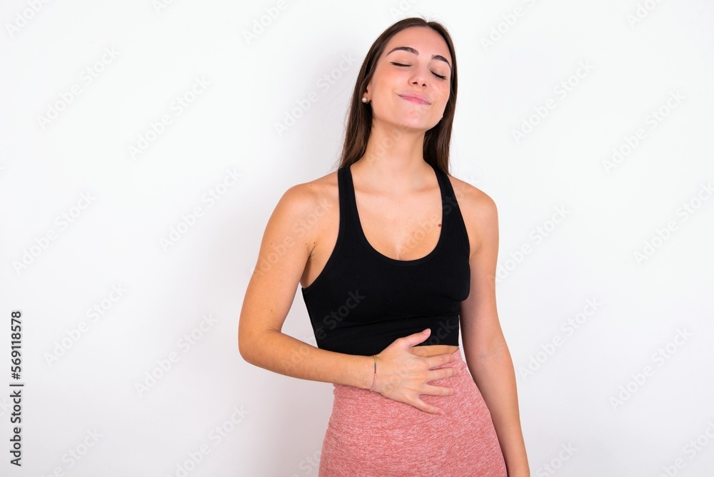 Satisfied smiling young woman wearing sportswear over white studio wall, keeps hands on belly, being in good mood after eating delicious supper, demonstrates she is full. Pleasant feeling in stomach. Satisfied smiling young woman wearing sportswear over white studio wall, keeps hands on belly, being in good mood after eating delicious supper, demonstrates she is full. Pleasant feeling in stomach.