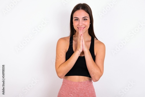 young woman wearing sportswear over white studio background keeps palms together, has pleased expression. Glad attractive male makes request, pleads for mercy. Hopeful young adult.