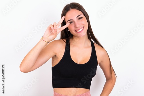 young woman wearing sportswear over white studio background making v-sign near eyes. Leisure, coquettish, celebration, and flirt concept.