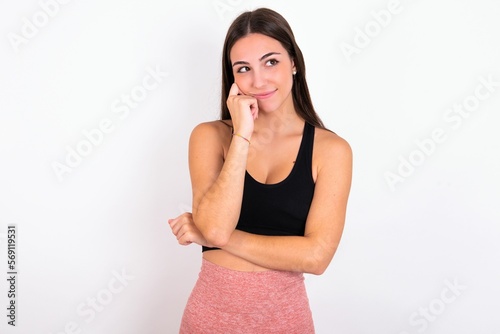 Face expressions and emotions. Thoughtful young woman wearing sportswear over white studio background holding hand under his head, having doubtful look.