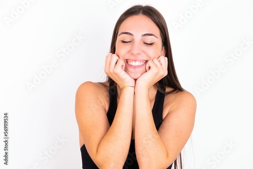 Portrait of young woman wearing sportswear over white studio background being overwhelmed, expressing excitement and happiness with closed eyes and hands near face.