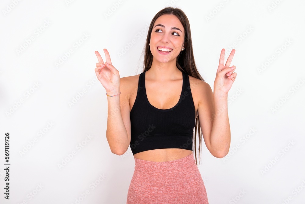 Fototapeta premium Isolated shot of cheerful young woman wearing sportswear over white studio background makes peace or victory sign with both hands, feels cool.