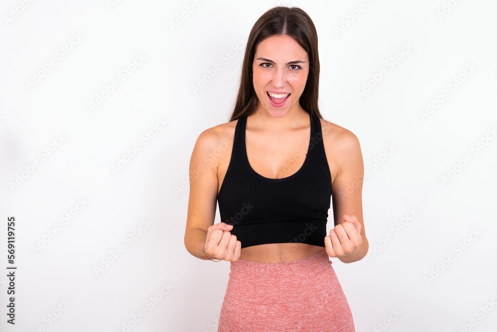 Joyful excited lucky young woman wearing sportswear over white studio background cheering, celebrating success, screaming yes with clenched fists Joyful excited lucky young woman wearing sportswear over white studio background cheering, celebrating success, screaming yes with clenched fists