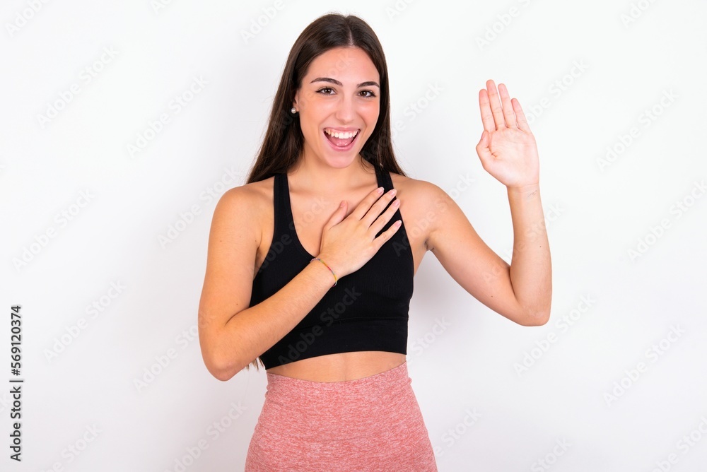 I swear, promise you not regret. Portrait of sincere young woman wearing sportswear over white studio wall raising one arm and hold hand on heart as give oath, telling truth, want you to believe.