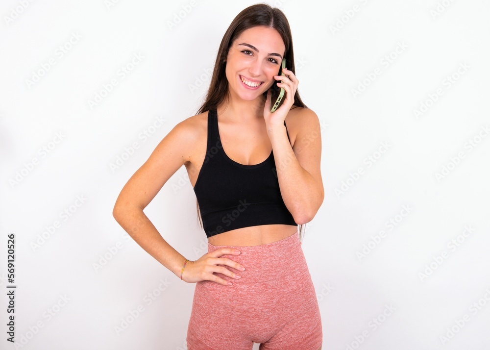 Portrait of successful joyful young woman wearing sportswear over white studio background talking on mobile phone with friend. Lifestyle and communication concept