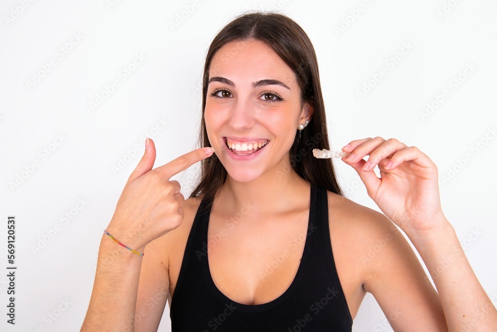 Φωτογραφία young woman wearing sportswear over white studio background holding an invisible aligner and pointing to her perfect straight teeth