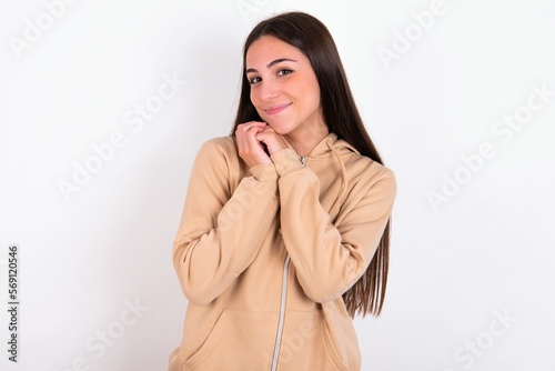 Charming serious young woman wearing beige sweater over white studio background keeps hands near face smiles tenderly at camera