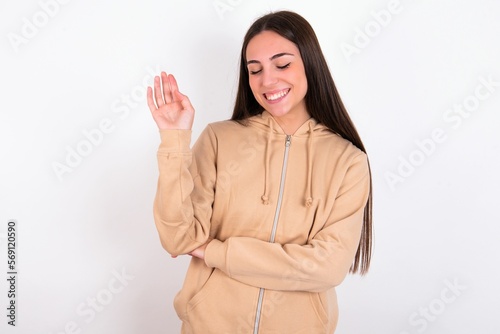 Overjoyed successful young woman wearing beige sweater over white studio background raises palm and closes eyes in joy being entertained by friends