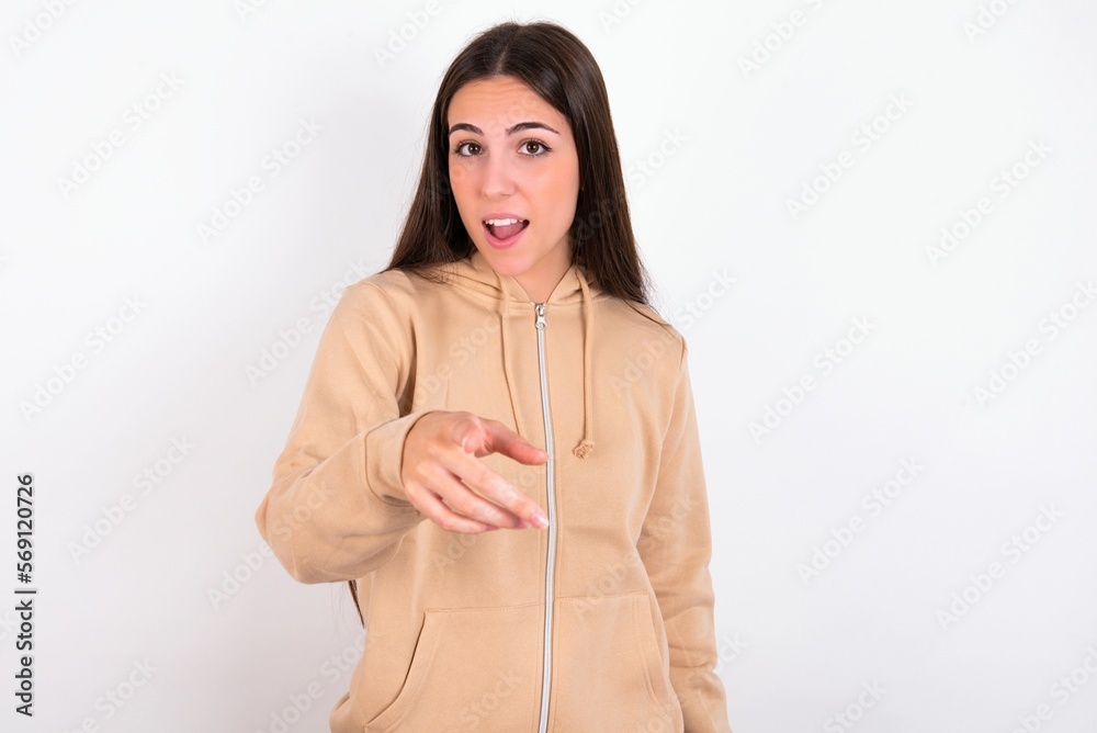 Shocked young woman wearing beige sweater over white studio background points at you with stunned expression Shocked young woman wearing beige sweater over white studio background points at you with stunned expression