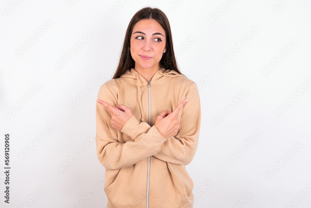 young woman wearing beige sweater over white studio background crosses arms and points at different sides hesitates between two items or variants. Needs help with decision