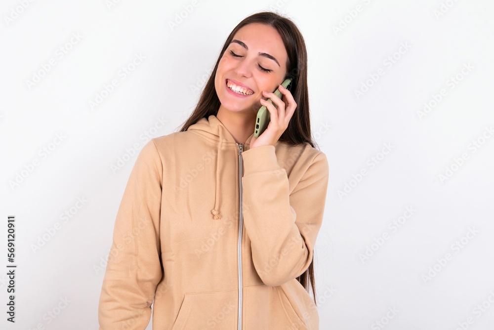 Funny young woman wearing beige sweater over white studio background laughs happily, has phone conversation, being amused by friend, closes eyes. Funny young woman wearing beige sweater over white studio background laughs happily, has phone conversation, being amused by friend, closes eyes.