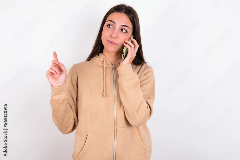 young woman wearing beige sweater over white studio background speaks on mobile phone spends free time indoors calls to friend. young woman wearing beige sweater over white studio background speaks on mobile phone spends free time indoors calls to friend.