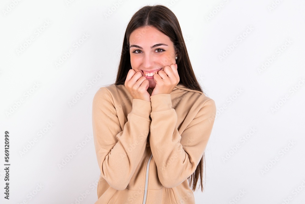 Satisfied young woman wearing beige sweater over white studio background touches chin with both hands, smiles pleasantly, rejoices good day with lover Satisfied young woman wearing beige sweater over white studio background touches chin with both hands, smiles pleasantly, rejoices good day with lover