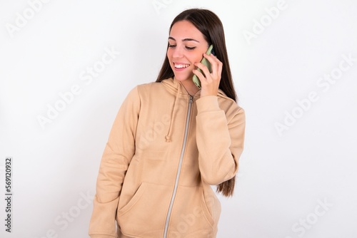 Pleasant looking happy young woman wearing beige sweater over white studio background has nice telephone conversation and looks aside, has nice mood and smiles positively while talks via cell phone