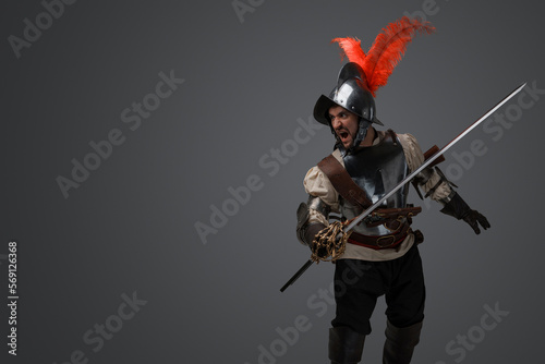Studio shot of isolated on gray background soldier with epee dressed in plate armor.