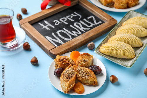 Chalkboard with text HAPPY NOWRUZ, treats and glass of tea on blue background, closeup