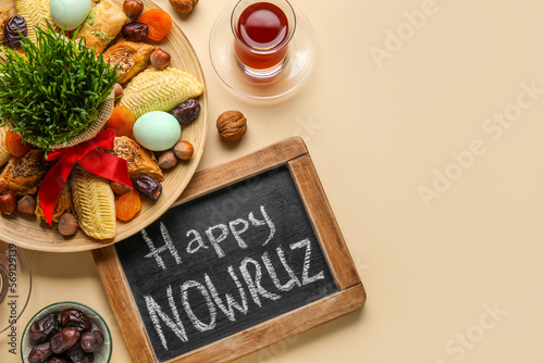 Chalkboard with text HAPPY NOWRUZ, treats, glass of tea and grass on beige background