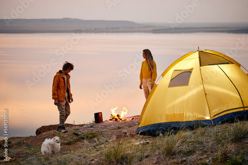Evening camping at lake. tourist couple, man and woman with dog next to campfire. Tourism, happy relations and active lifestyle concept. caucasian married couple in sportswear relaxing, enjoying rest