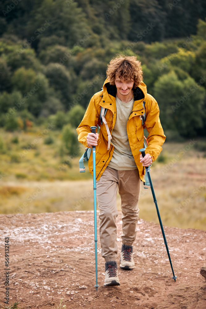 Healthy hiker man in nature hike. Athlete young male hiking happy with Trekking pole stands on meadow. Background beautiful mountains with green trees. curly caucasian traveller in yellow coat