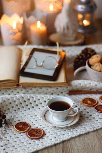 Cup of tea  books  tablet  bowl of cookies  various spices  pine cones and lit candles. Hygge at home. Selective focus.