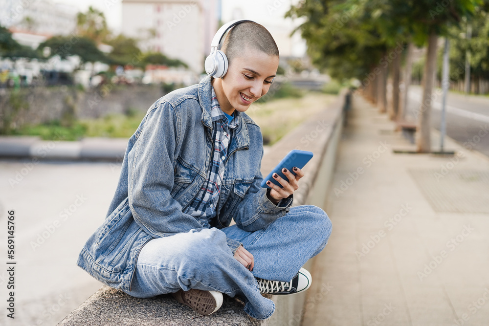 Young bald woman listening music playlist while using mobile phone outdoor - Focus on face