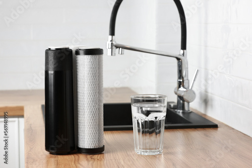 A glass of clean fresh water and set of filter cartridges on wooden table in a kitchen interior. Installation of reverse osmosis water purification system. Concept Household filtration system. photo