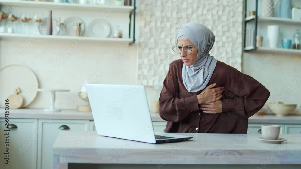 Young Muslim woman with severe heartache feels bad A female in a hijab suffers from chest pain while sitting at home working on a laptop computer alone indoor having heart attack disease Young Muslim woman with severe heartache feels bad A female in a hijab suffers from chest pain while sitting at home working on a laptop computer alone indoor having heart attack disease