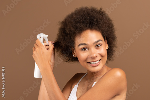 Smiling young african american curly female with perfect skin in white top applying spray on her hair