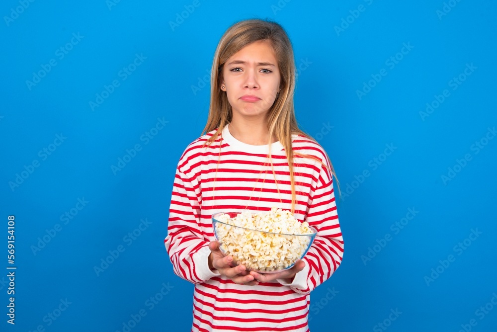 Displeased upset caucasian teenager girl wearing striped T-shirt over blue background frowns face as going to cry, being discontent and unhappy as can't achieve goals,  eating popcorn