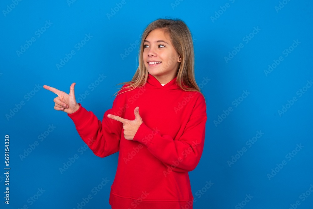 Optimistic caucasian teenager girl wearing red sweater over blue background points with both hands and looking at empty space. Optimistic caucasian teenager girl wearing red sweater over blue background points with both hands and looking at empty space.