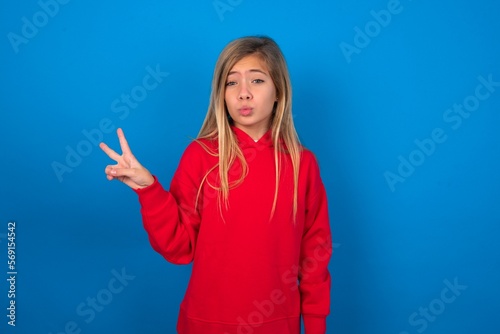 caucasian teenager girl wearing red sweater over blue background makes peace gesture keeps lips folded shows v sign. Body language concept