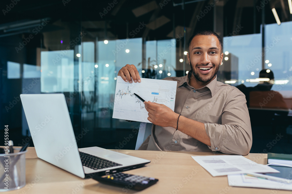 Portrait of young successful financier paperwork inside office, man smiling and looking at camera shows graph with positive dynamics of economic profits, businessman at work with laptop.