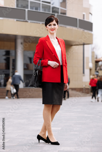 Smiling business woman dressed red jacket, black skirt with leather business bag standing outdoor near corporate office building, hand on the hip. Confident caucasian female business on city street.