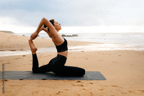 Sporty woman practising yoga on fitness mat on the beach by seaside, enjoying training outdoors, side view, copy space