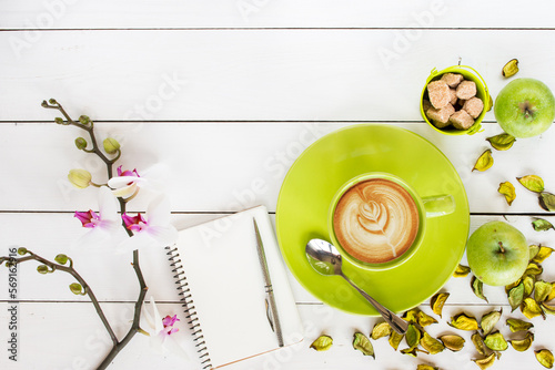 cappuccino coffee in green cup,  orchid flower, notebook and silver pen,  green dry flower decor scattered on white painted wooden table, top view