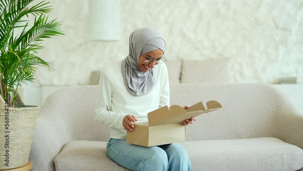 Young Muslim woman in hijab received a parcel satisfied with shopping in online internet store sitting on sofa at home Happy customer rejoice with purchase order while opening carton box indoors Young Muslim woman in hijab received a parcel satisfied with shopping in online internet store sitting on sofa at home Happy customer rejoice with purchase order while opening carton box indoors