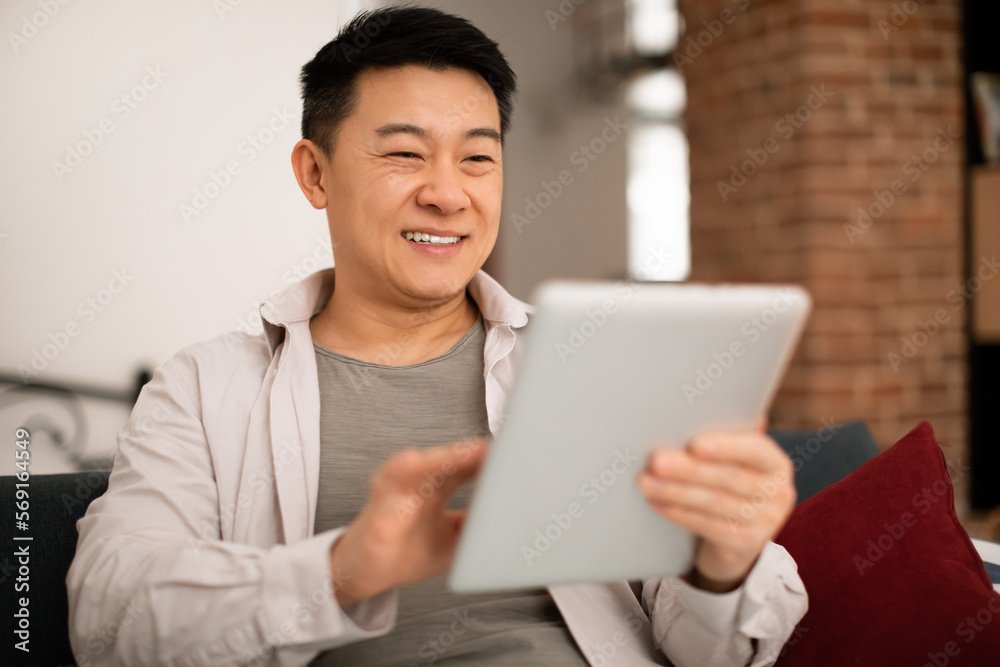 Technologies for leisure. Happy asian man using digital tablet at home, resting with modern gadget on sofa, closeup Technologies for leisure. Happy asian man using digital tablet at home, resting with modern gadget on sofa, closeup