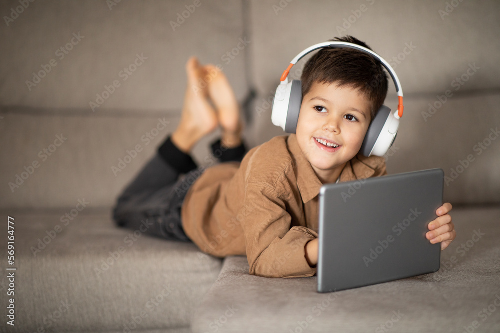 Smiling caucasian little kid in headphones enjoys free time, watching video, playing game on tablet Smiling caucasian little kid in headphones enjoys free time, watching video, playing game on tablet