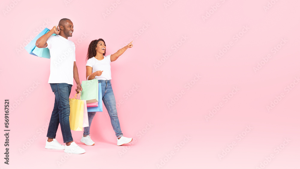 Naklejka premium Seasonal discounts. Black spouses walking with shopping bags and pointing at copy space on pink background, full length