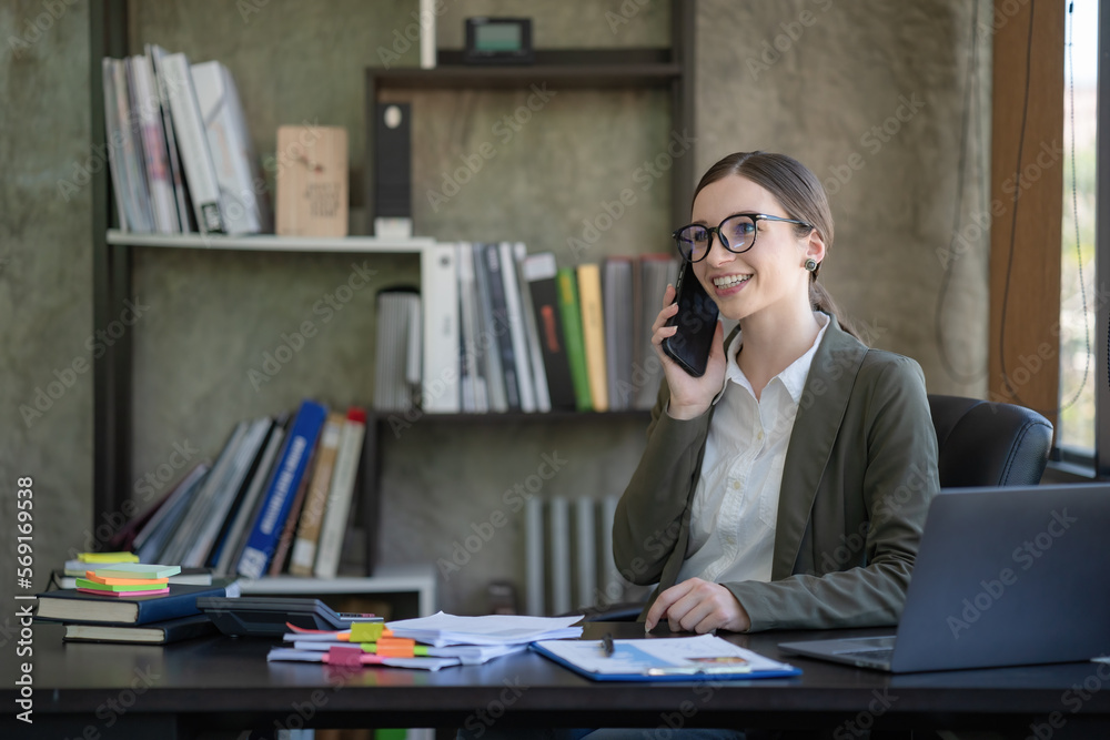 A happy young woman talking on her mobile phone and smiling while sitting at her working place in the office. A happy young woman talking on her mobile phone and smiling while sitting at her working place in the office.