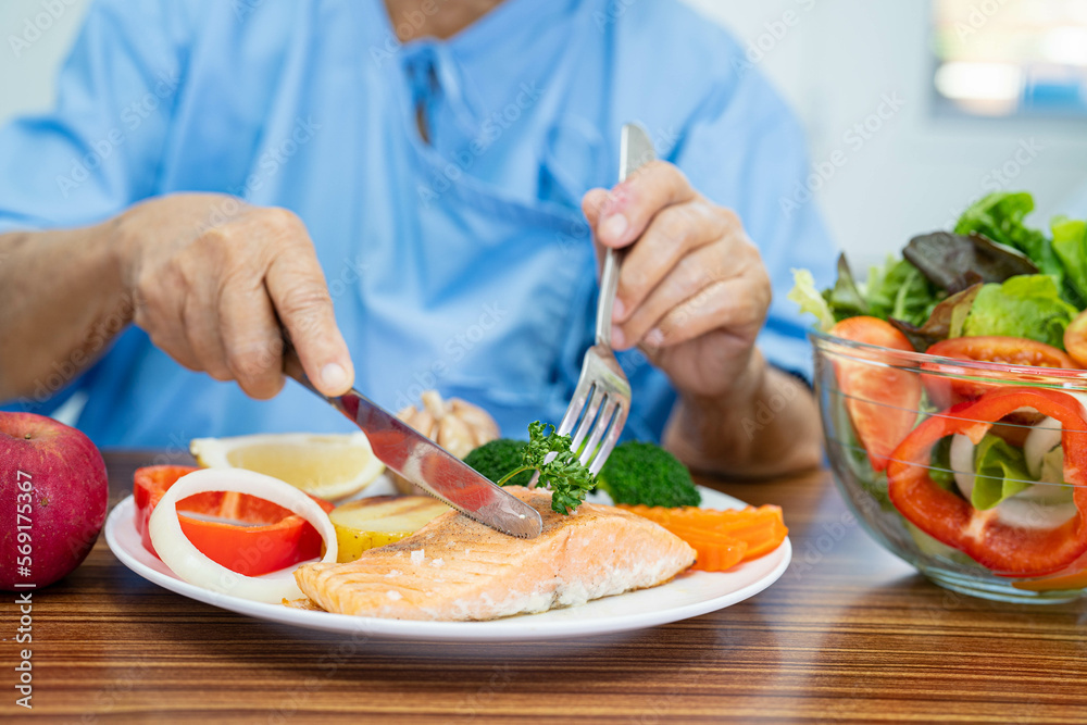 Asian senior or elderly old lady woman patient eating Salmon steak breakfast with vegetable healthy food while sitting and hungry on bed in hospital.