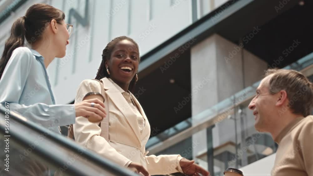 Three cheerful young diverse friends going down escalator in modern shopping centre and having conversation Three cheerful young diverse friends going down escalator in modern shopping centre and having conversation