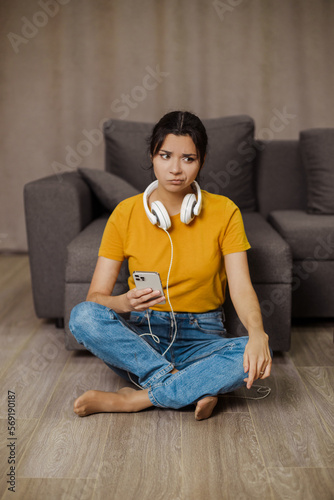 An unhappy Indian or arabian girl sits on the floor at home and can't listen to music, her phone is discharged
