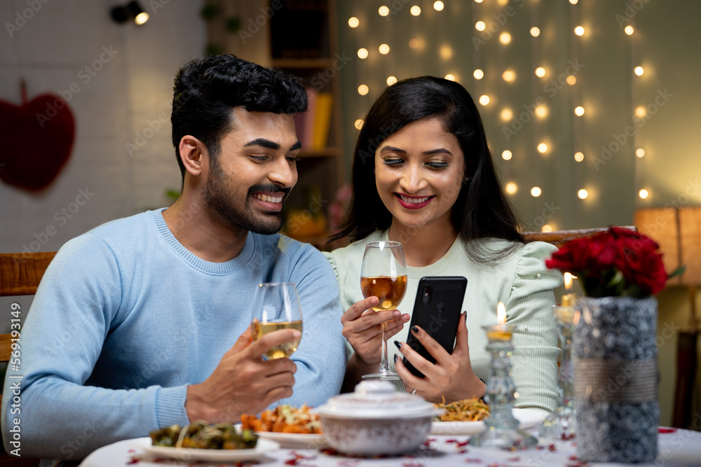 Happy couple using mobile phone while having drinks during romantic candlelight dinner - concept of wedding anniversary celebration, bonding and dating application