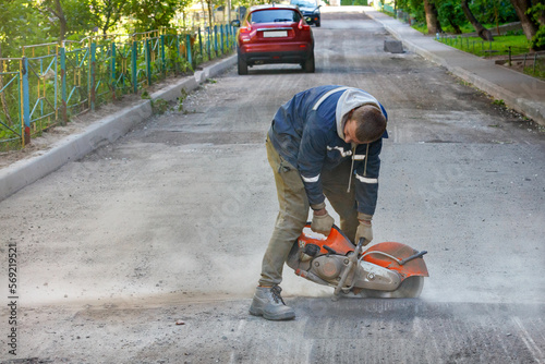 A worker cuts worn-out asphalt with a petrol cutter in a cloud of dust against the backdrop of a road being repaired.
