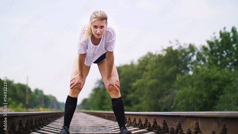 Beautiful sexy athletic young blond woman in top and shorts, running, jogging, Stops, rests, breathes heavily, is tired, looks at the camera. On the railway, in the summer.