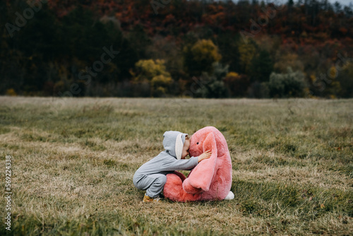 Little child wearing a bunny costume, playing with a big pink plush bunny toy, hugging it, outdoors, in an open field.