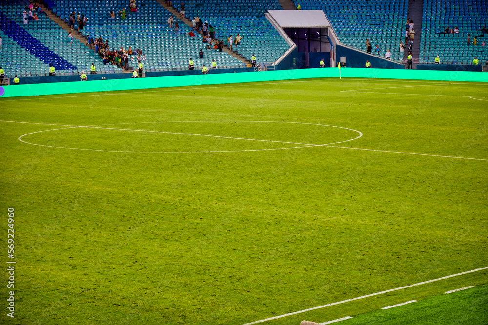 Obraz premium High stands of the football stadium are gradually filled with fans and supporters. Large green field for playing football prepared before the match